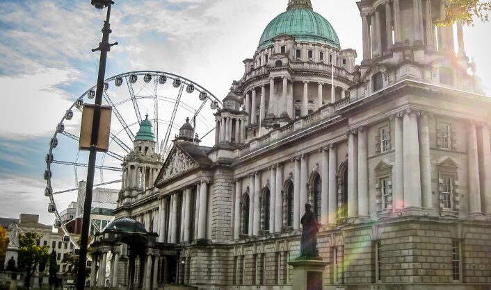 Belfast City Hall with its green copper dome, with a Ferris wheel behind it and soft sunlight lens flare in the foreground.
