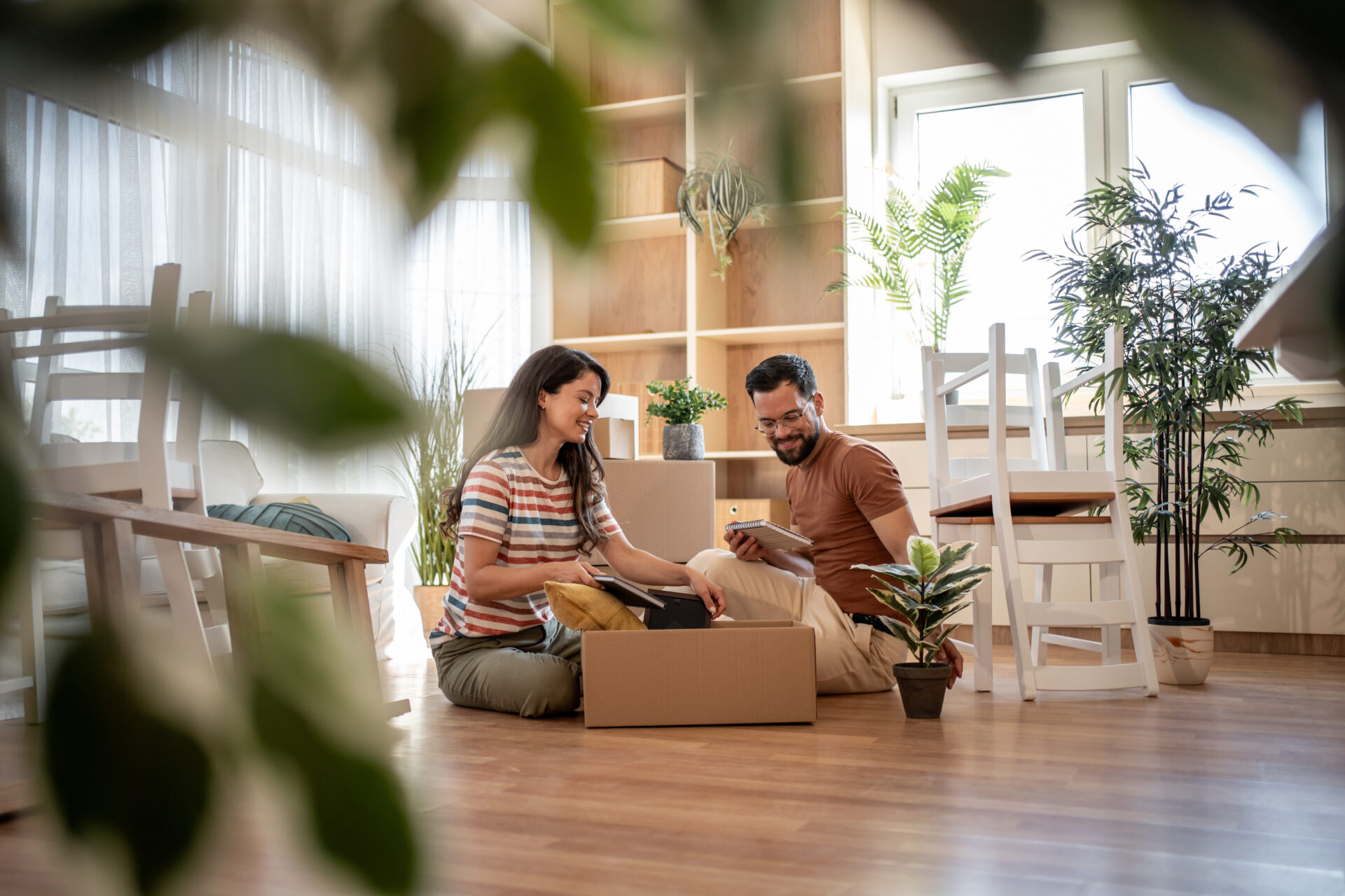 Young smiling couple unpacking belongings from cardboard boxes after moving into new home, sitting on floor and using digital tablet for faster organization