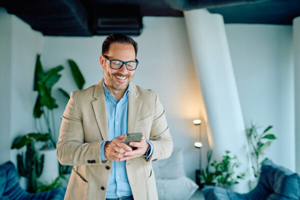 Smiling businessman using cell phone, connecting with work or personal messages, enjoying modern technology inside an office lounge